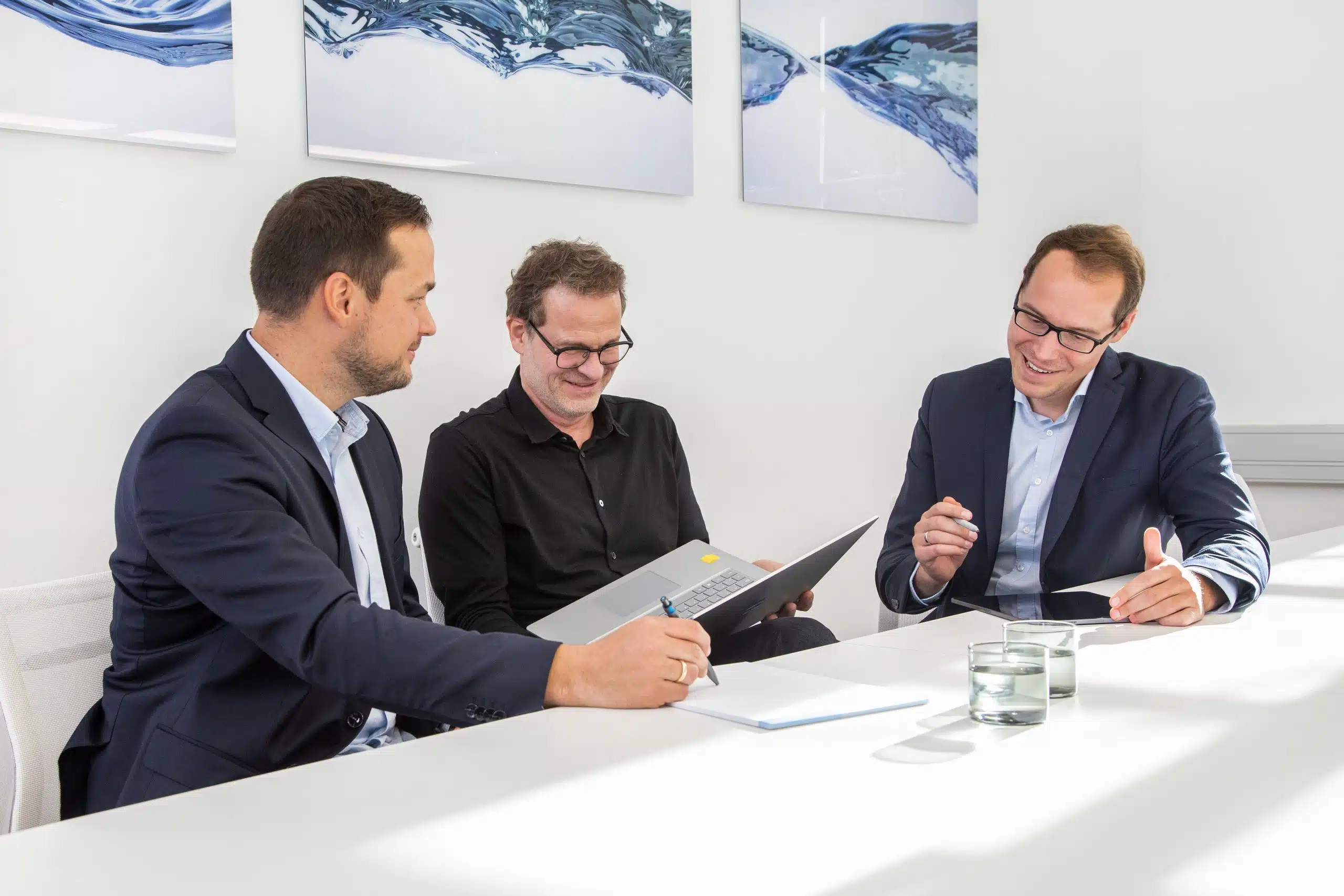 Drei Steuerberater, Frank Bott, Florian Brauers und Robert Kopiec sitzen am Konferenztisch in einem hellen Büro. Sie arbeiten am Laptop, beraten sich und lachen herzlich. Im Hintergrund ist moderne Kunst zu sehen.