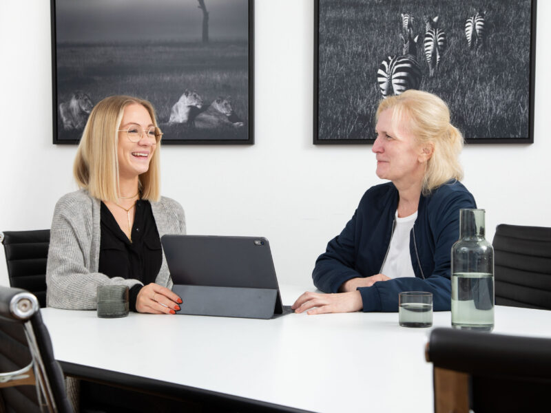 zwei Steuerfachangestellte, Anja Michel und Corinna Lange lachen, sitzen am Tisch im Buero und arbeiten am Laptop. Im Hintergrund ist moderne Schwarzweiß Fotografiekunst zu sehen