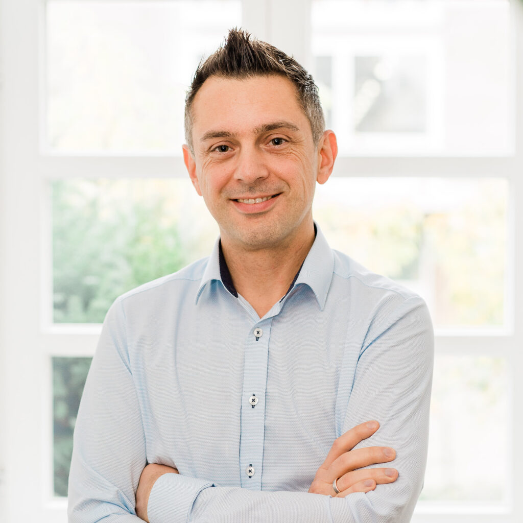 Portrait eines lächelnden Mitarbeiters, Gordon, der vor einem hellen Altbau-Sprossenfenster der Kanzlei steht. Der Hintergrund ist unscharf. Er hat die Arme verschränkt. Er trägt ein hellblaues Hemd und kurze Haare. Sein Pony ist mit Gel nach oben gestylt.