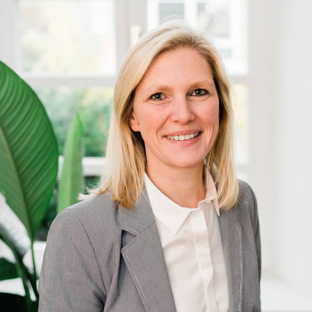 Eine laechelnde Mitarbeiterin, Sylvia, steht vor einem Altbau-Sprossenfenste und einer Gruenpflanze mit großen gruenen Blaettern