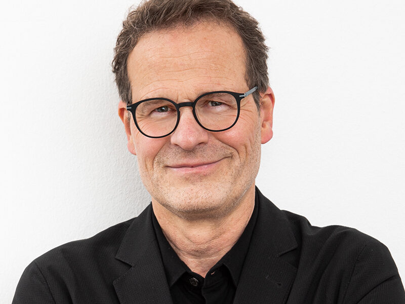Brustportrait von Frank Bott Steuerberater, mit Brille und schwarzem Hemd steht vor weißer Wand und lächelt