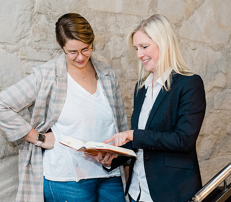 Zwei Mitarbeiterinnen im Flur der Steuerberaterkanzlei schauen in ein Buch und lächeln