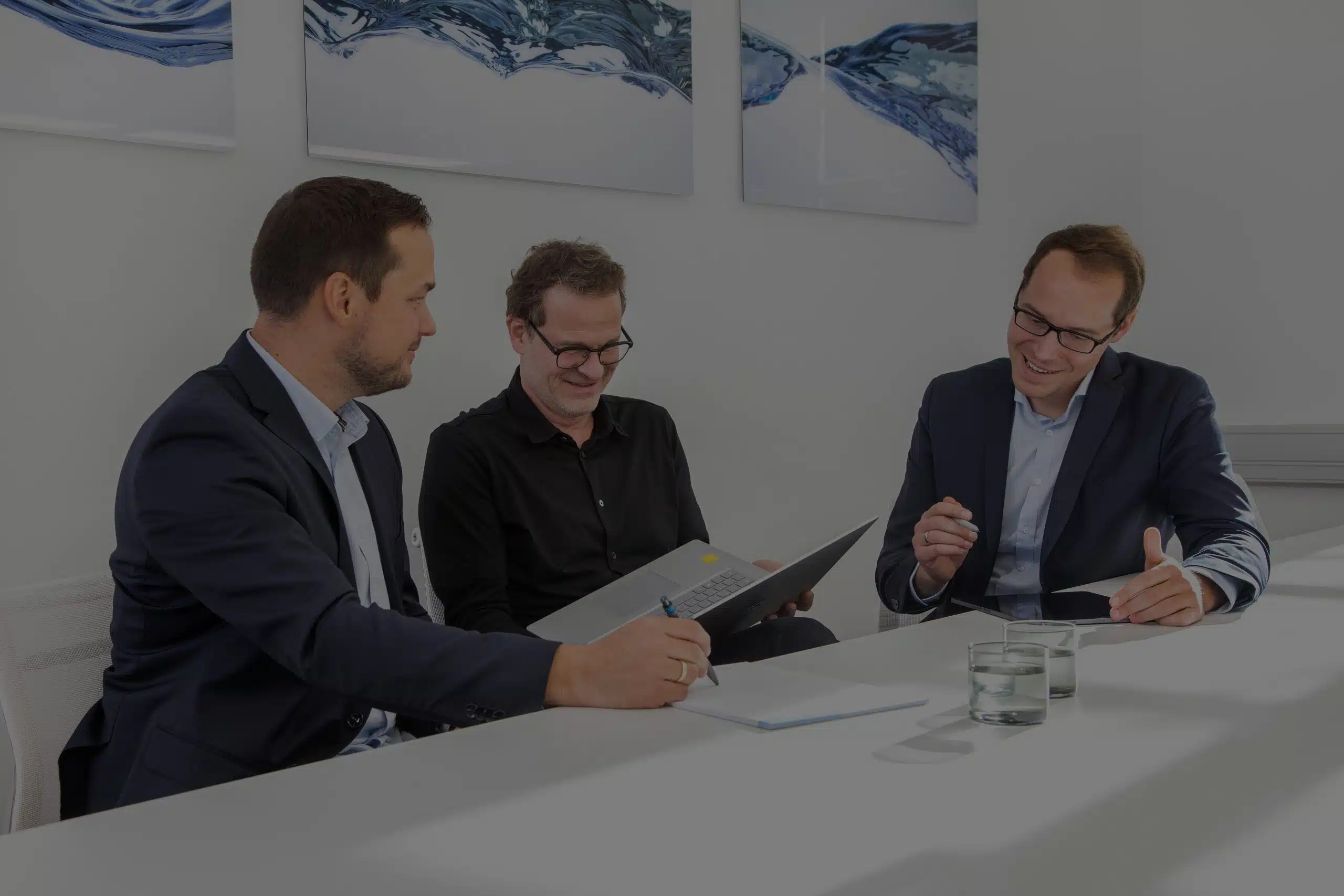 Frank Bott, Florian Brauers und Robert Kopiec, die drei Steuerberater der Kanzlei sitzen am Konferenztisch und arbeiten am Laptop, beraten sich und lachen herzlich. Im Hintergrund moderne Kunst. Das Bild hat eine schwarze Überlagerung von 60 Prozent.