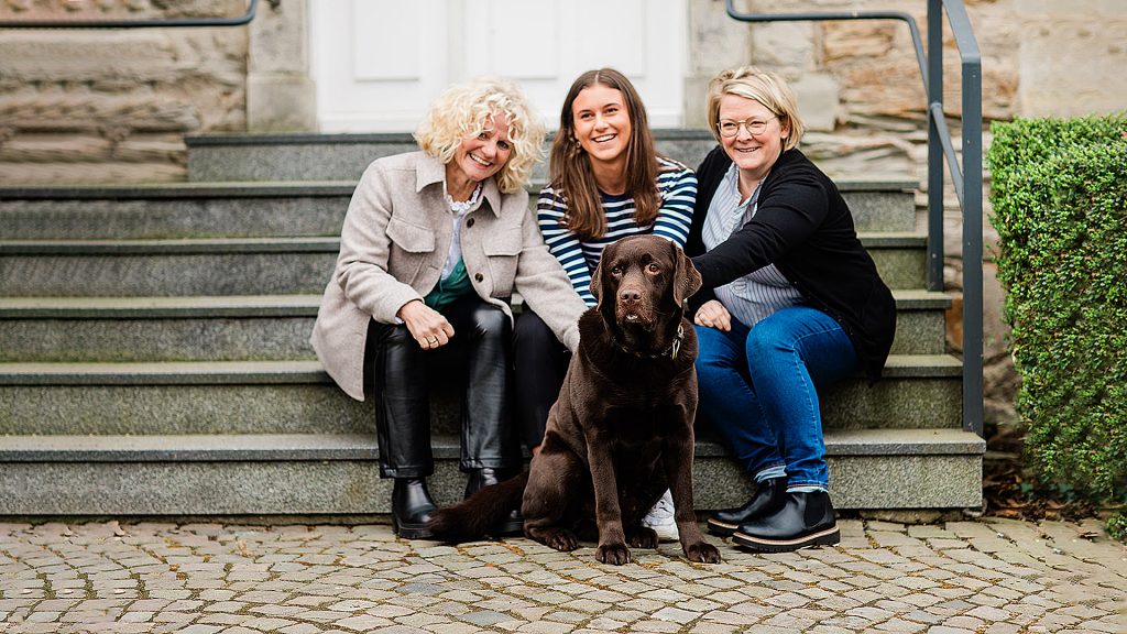 Drei Frauen sitzen auf der Treppe vor der Kanzlei und streicheln einen braunen Labrador
