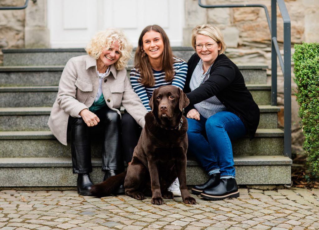 Drei Frauen sitzen auf der Treppe vor der Kanzlei und streicheln einen braunen Labrador